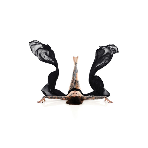 Artistic high-key photo of a tattooed woman lying on her back with legs gracefully raised and sheer black fabric flowing from her arms, symbolizing empowerment and self-expression in Nuovo's Self-Love collection