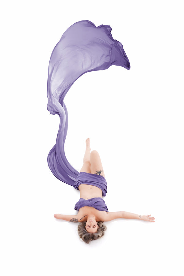 Woman embracing self-love in a high-key photography style, lying with arms spread and a flowy lavender fabric creating a wave overhead, symbolizing freedom and confidence