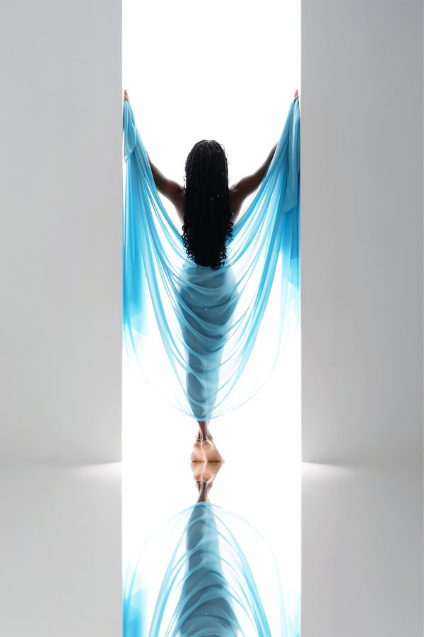 An empowering high-key portrait from Nuovo's Self-Love collection, featuring a person enveloped in a cascading sky-blue fabric, with arms wide open and a reflection beneath, embodying a moment of self-embrace and acceptance.